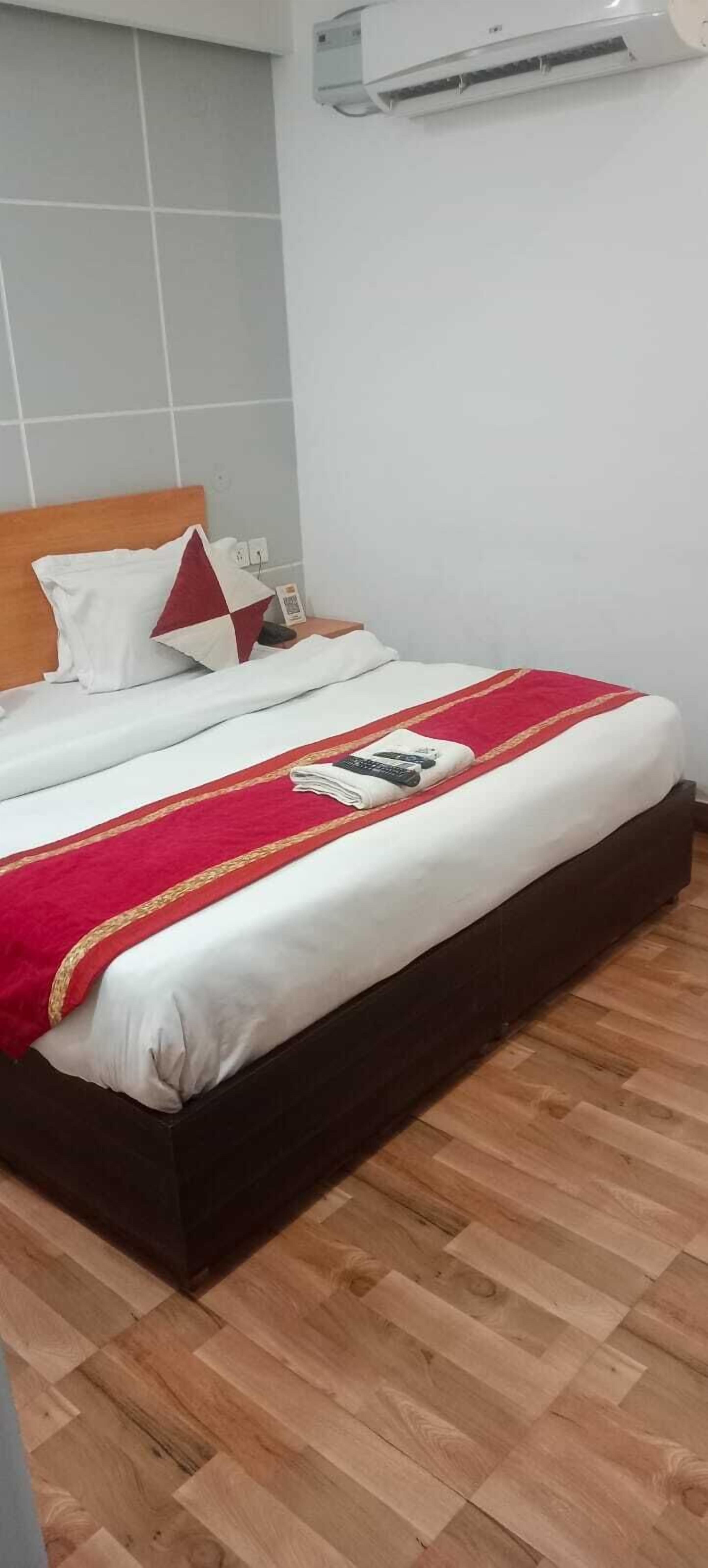 A hotel room with a wooden floor, a bed with white bedding and a red blanket, a wooden headboard, and an air conditioning unit on the wall.