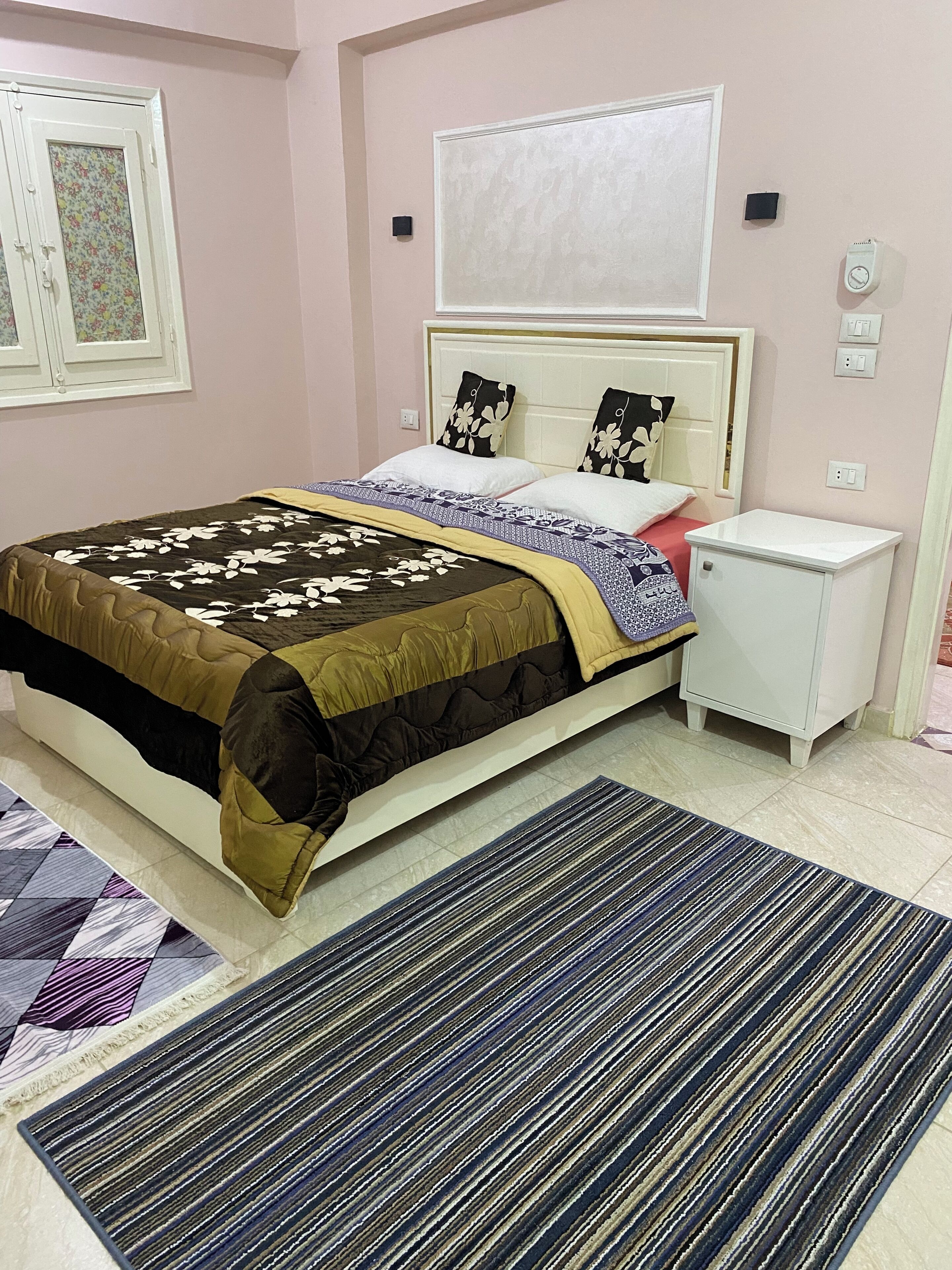 Quarto com cama, mesa de cabeceira e janela com padrão floral.