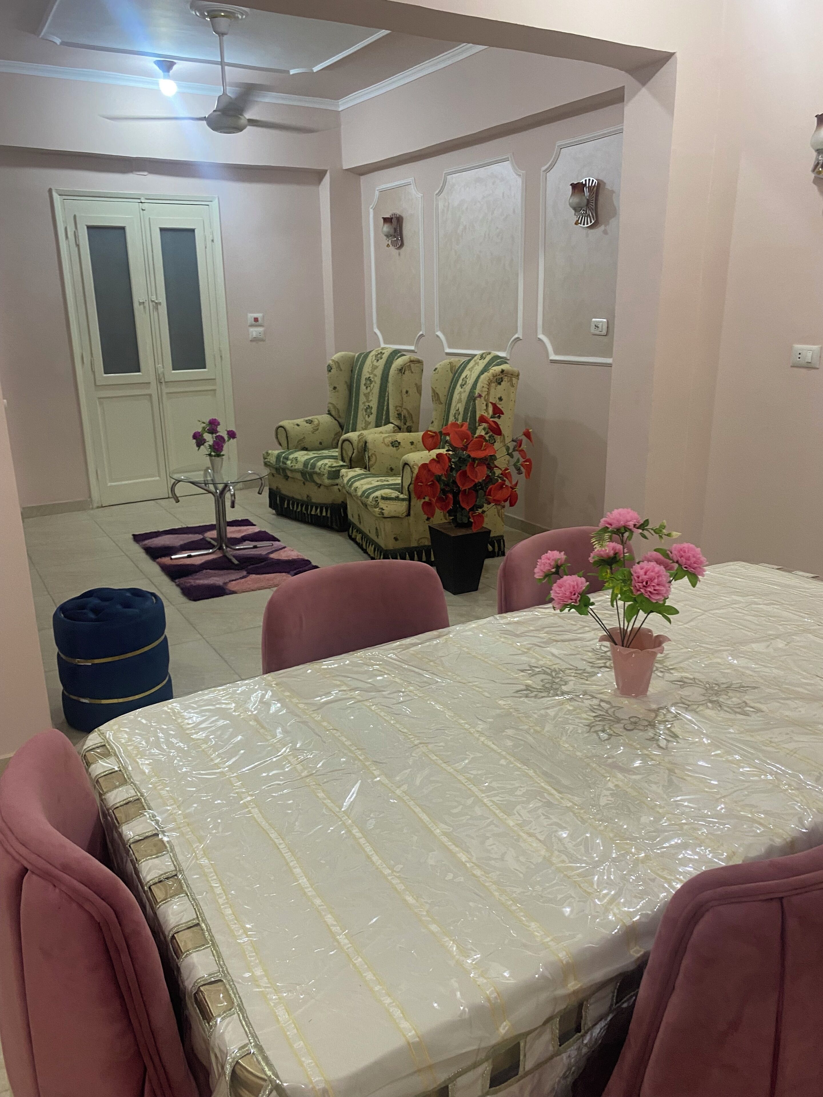 Un coin repas avec une table recouverte d’une feuille de plastique, entourée de chaises roses. On y trouve deux fauteuils à motifs floraux, un pouf bleu et un vase avec des fleurs rouges posé sur la table.