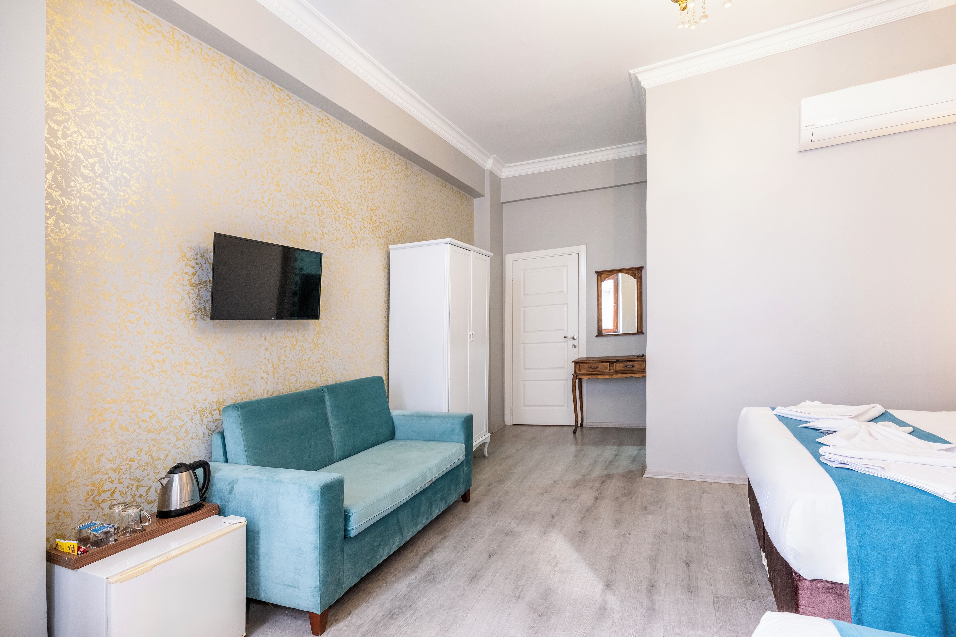 A hotel room with a teal sofa, a small white cabinet, a television mounted on the wall, a bed with a blue blanket, and a wooden floor.