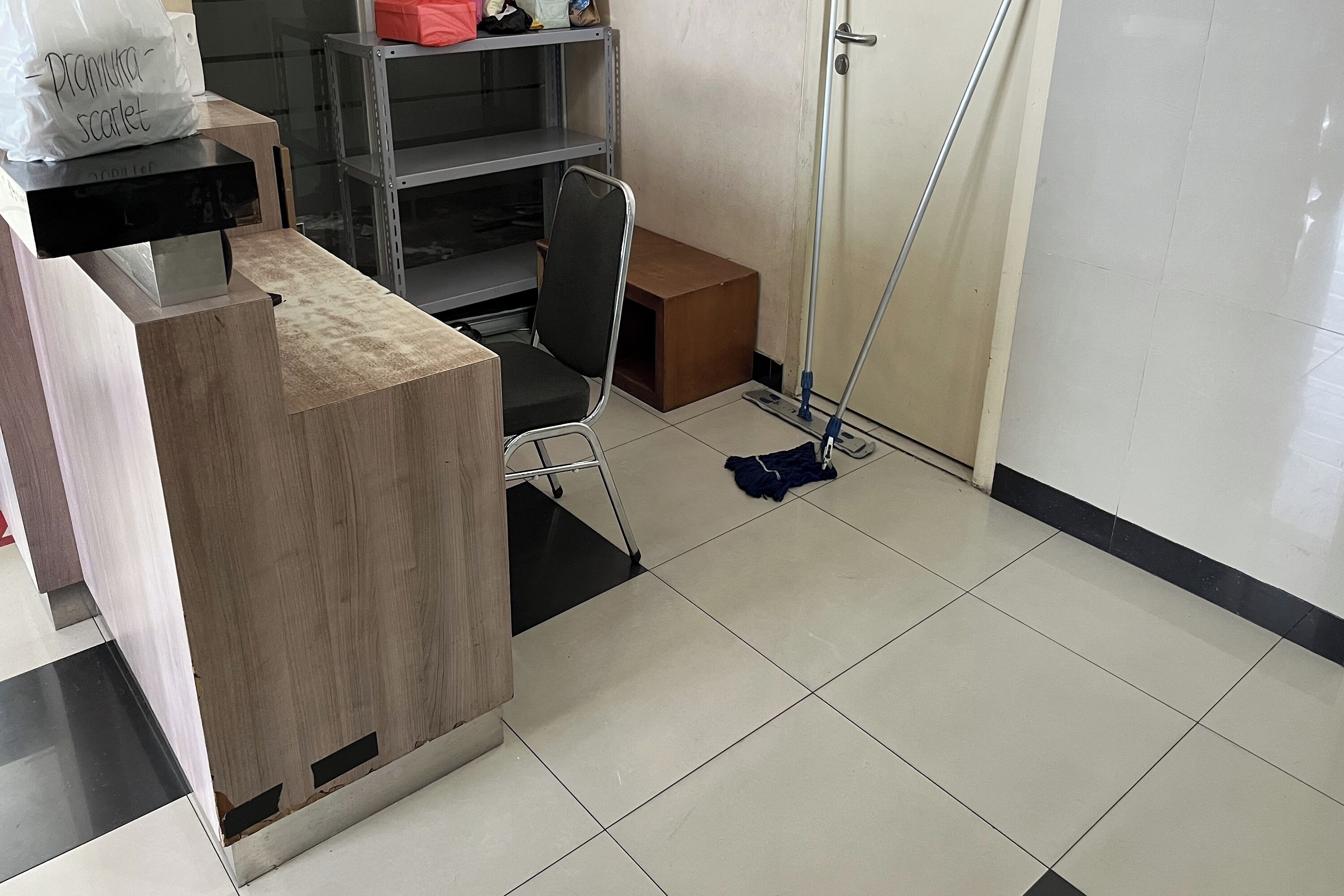 Une salle d’attente avec un bureau en bois, un fauteuil de bureau noir, une étagère en métal et une serpillière posée sur le sol.