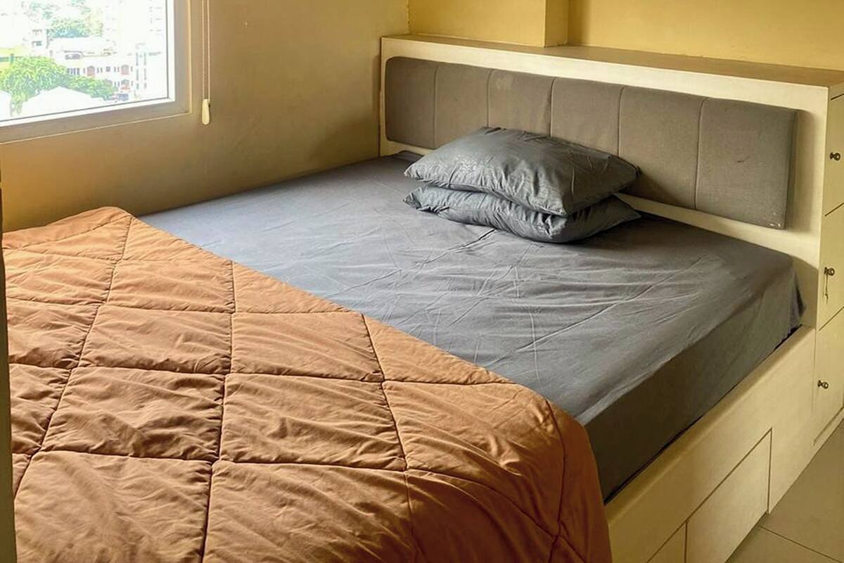 A bed with a quilted brown cover and a grey mattress, a grey pillow, and a headboard with a light-colored panel.