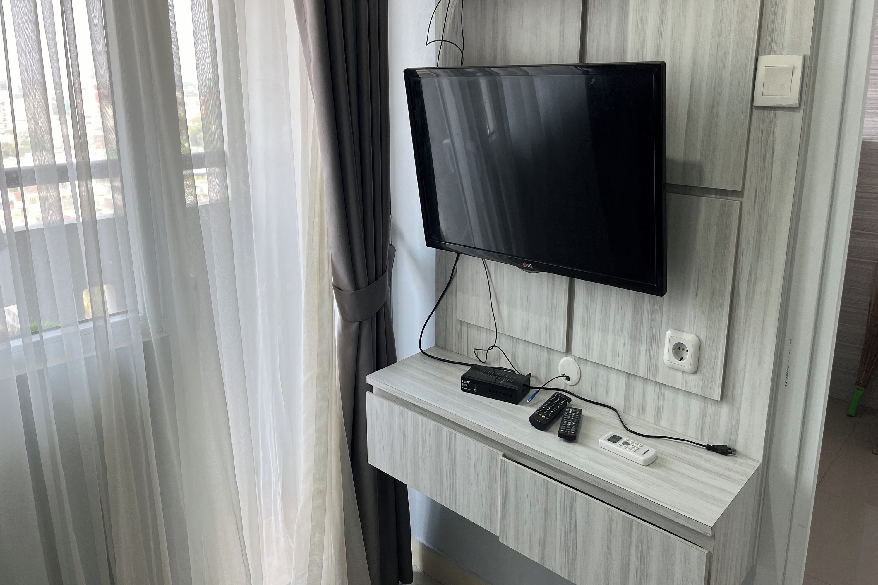 A wall-mounted TV with a control panel, remote controls, and a power strip on a light-colored wooden panel wall.