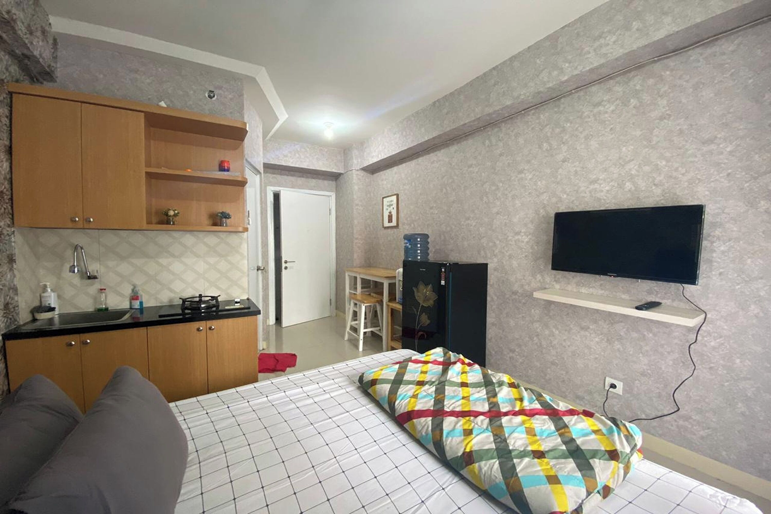 A compact hotel room with a bed, a small kitchenette, a TV mounted on the wall, and a door leading to a bathroom.