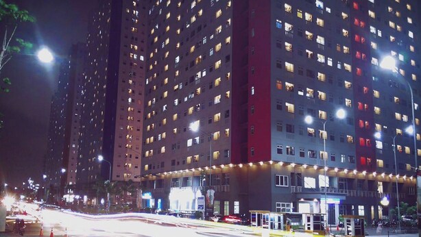 A high-rise building with multiple floors, illuminated windows, and a street with traffic lights and a bus stop. - Green Pramuka By Uchi Property (Jakarta)