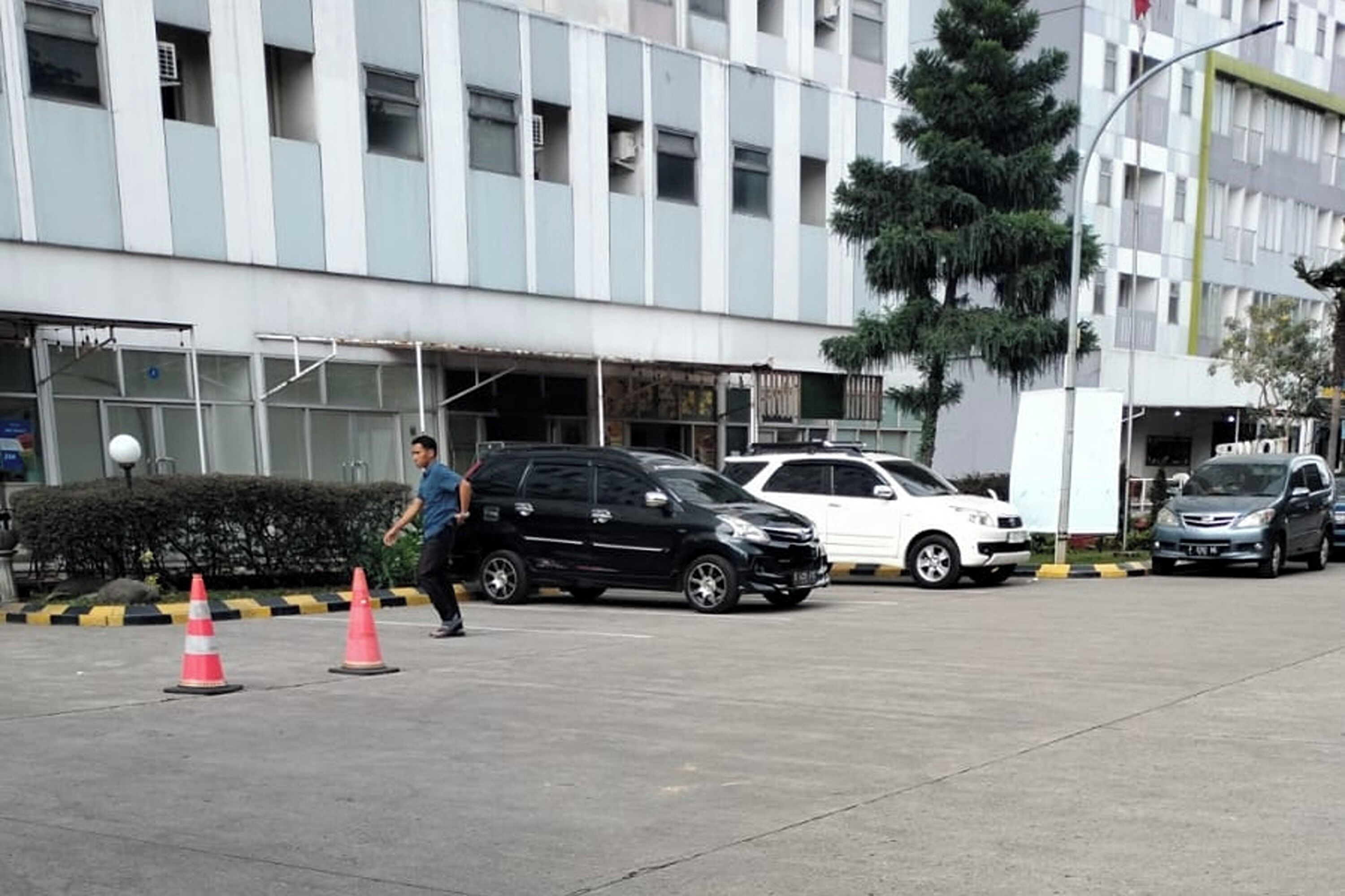 Un parking avec plusieurs voitures, un bâtiment doté d’une façade moderne et une personne qui marche.