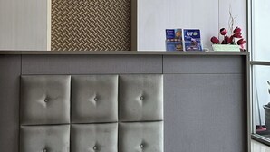 Egy modern fogadótér, szürke, párnázott fejrészessel, szövetből készült, mintás falfelülettel, valamint egy polccal, melyen folyóiratok és virágok találhatók.