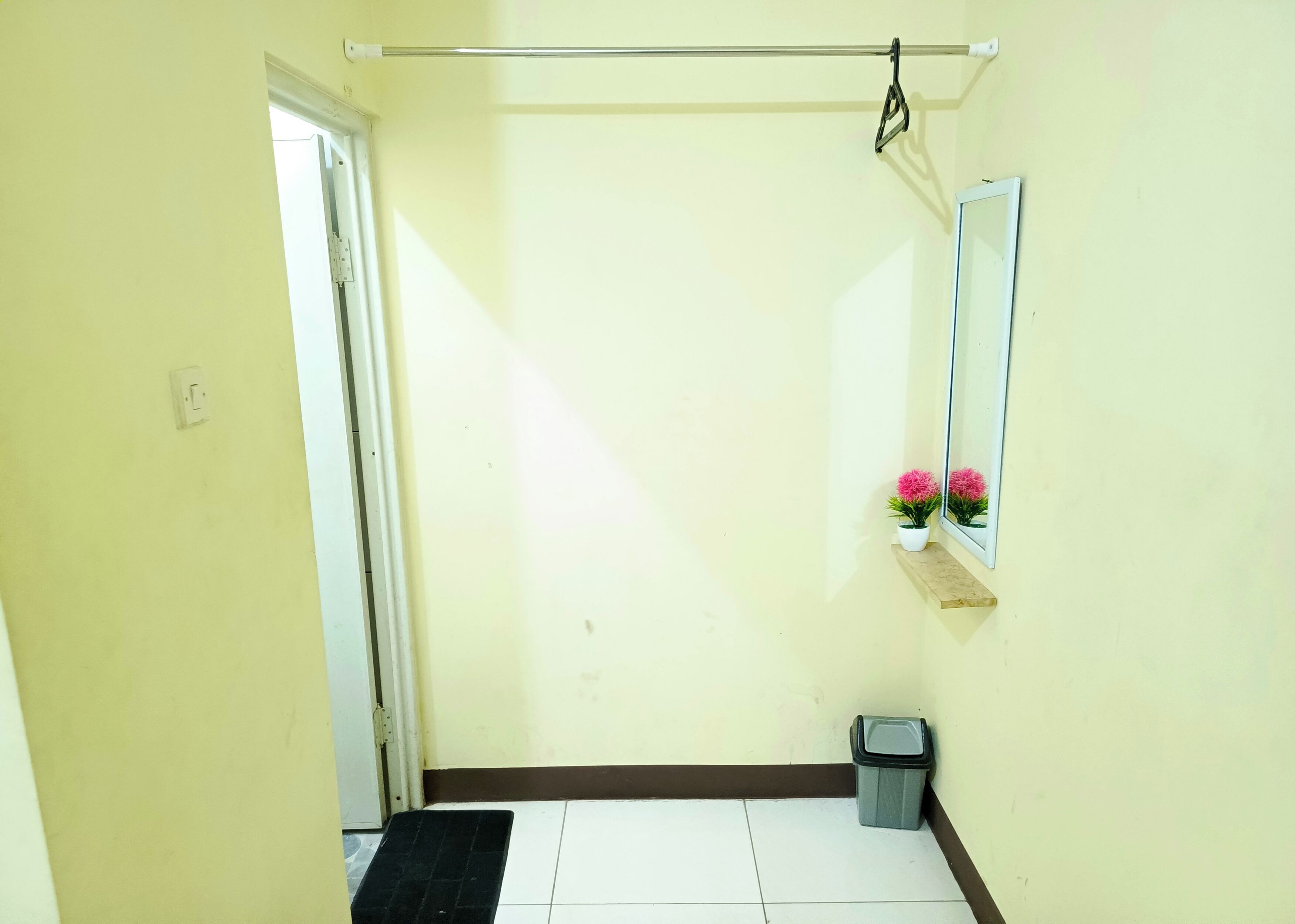 A narrow hallway with a door on the left, a trash bin on the right, and a small shelf with pink flowers.