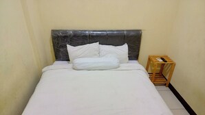 Ein Einzelbett mit weißer Bettwäsche und Kissen, ein Nachttisch aus Holz mit Fernbedienung und ein kleiner Holzschemel.