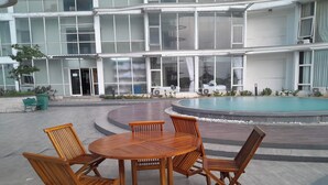 A wooden table and chairs set outdoors in front of a building with large windows and a swimming pool.