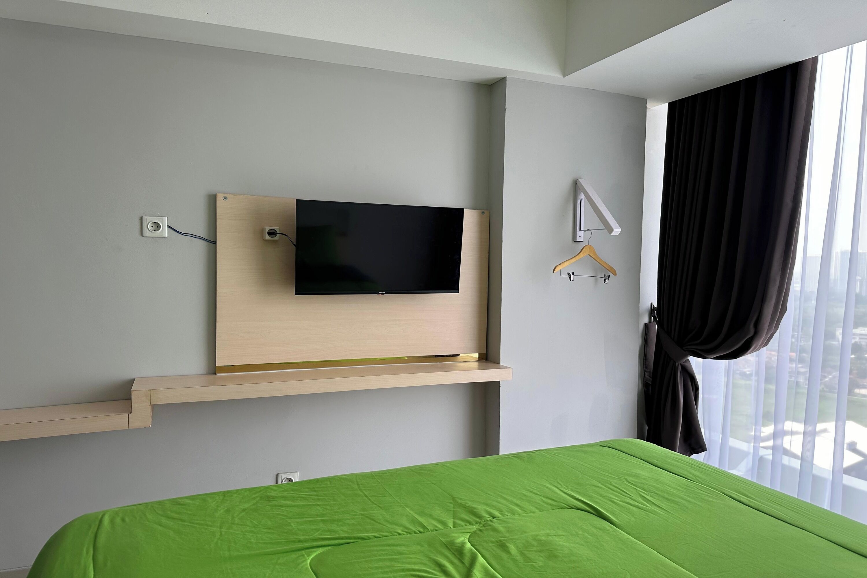 Una habitación de hotel con una cama, un televisor montado, una ventana con cortinas y un estante de madera.