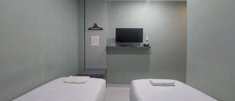 A small, clean room with two single beds, a flat-screen TV mounted on the wall, and a small table with a remote control.
