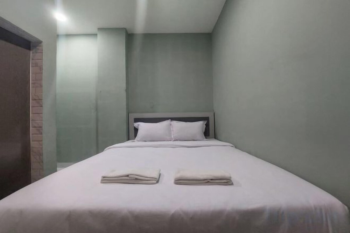 A single bed with white linens and pillows, a wooden door to the left, and a plain wall with a light switch above it.