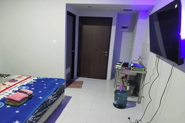 A bedroom with a bed, a TV mounted on the wall, a water dispenser, and a small table with items on it. - Riverdale Cikarang By Veto Rooms (Cikarang)