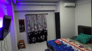 A bedroom with a bed, a television, an air conditioner, a window with floral curtains, and a small stool. - Riverdale Cikarang By Veto Rooms (Cikarang)