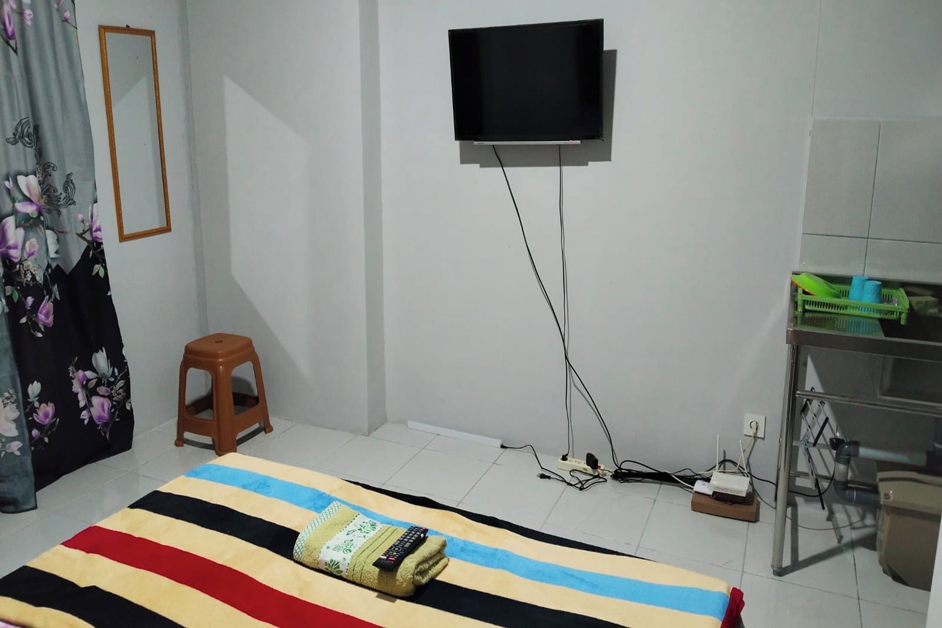 A bedroom with a bed, a television mounted on the wall, a stool, and a desk with a mirror.