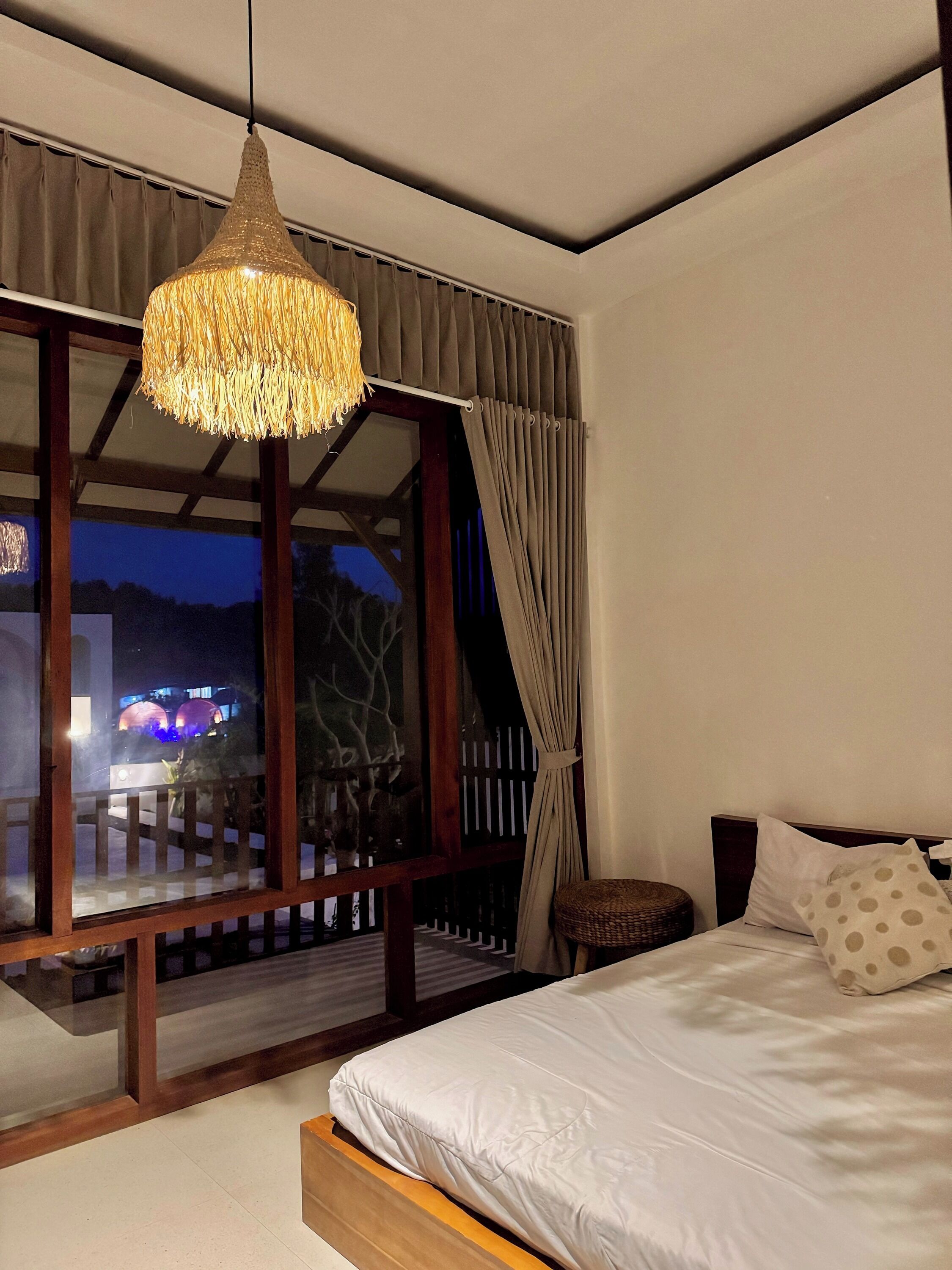 A bedroom with a large bed, a hanging light fixture, a view of the outside, and a balcony with a railing.