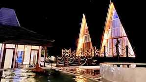 A nighttime view of a resort with two illuminated triangular buildings, a swimming pool, and a covered area with a view of the sea. - Volcano Side Kintamani Bali (Kintamani)