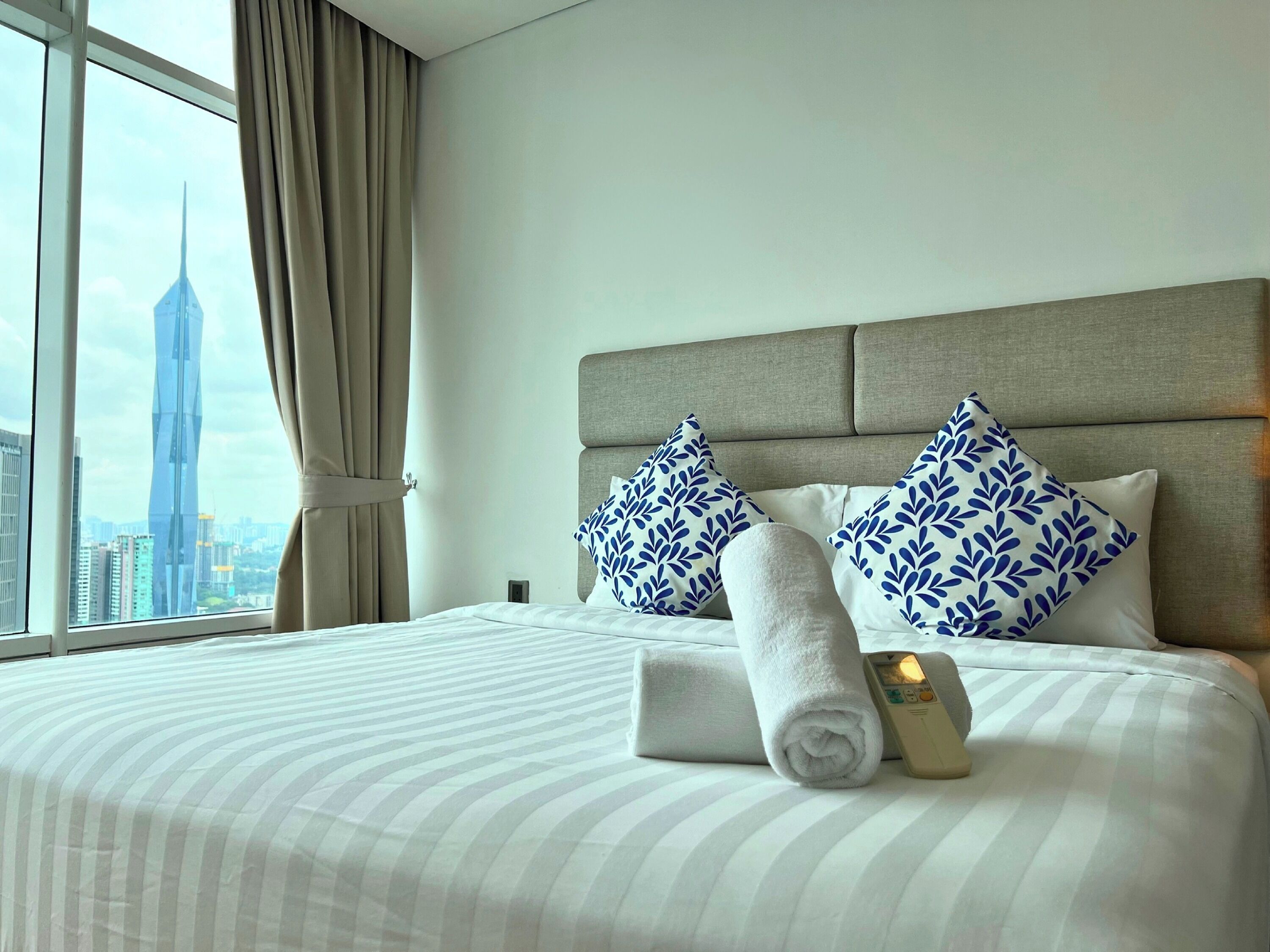 A hotel room with a large window, a bed with striped bedding, two pillows with a leaf pattern, and a rolled towel.