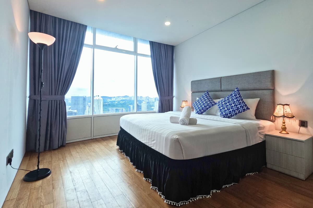 A modern hotel room with a large bed, a nightstand, a lamp, and a view of the city through large windows.