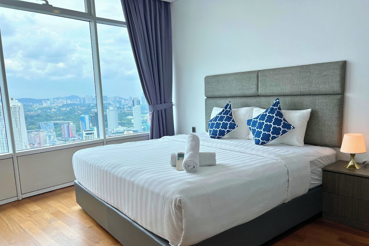 A modern hotel room with a large bed, grey headboard, and a view of the cityscape through large windows.