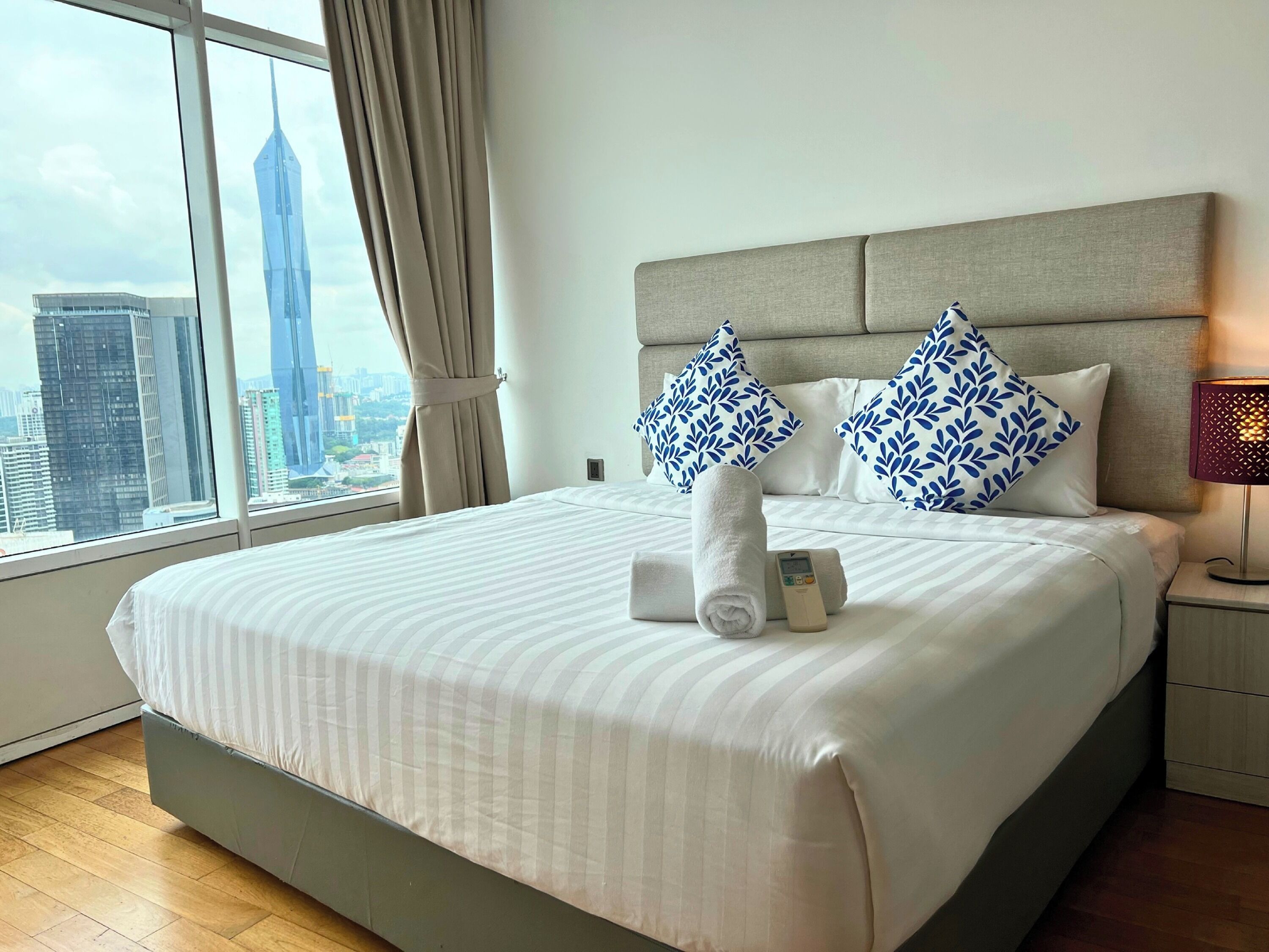 A hotel room with a large bed, a nightstand, a lamp, and a view of a cityscape through a window.
