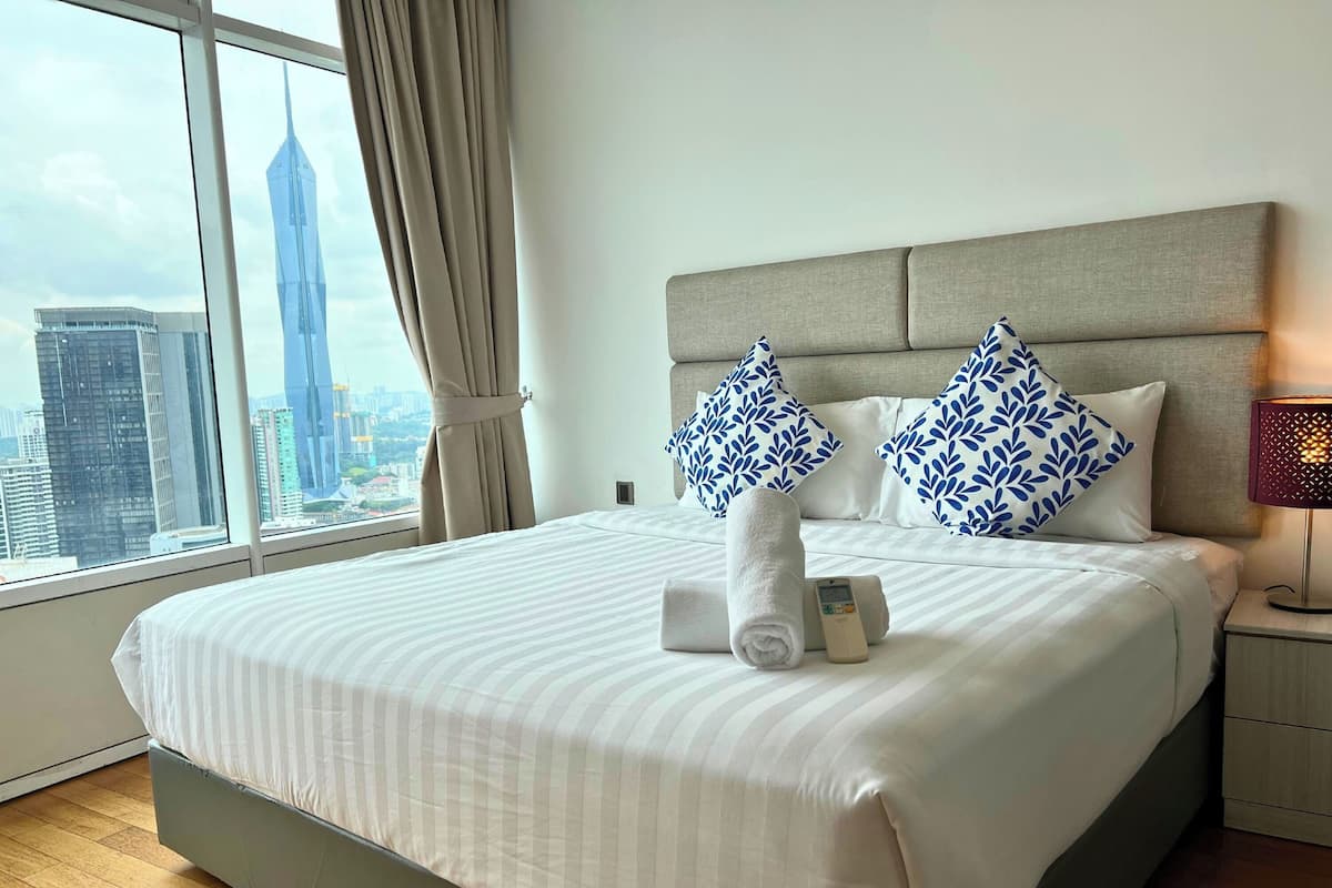A hotel room with a large bed, a nightstand, a lamp, and a view of a cityscape through a window.