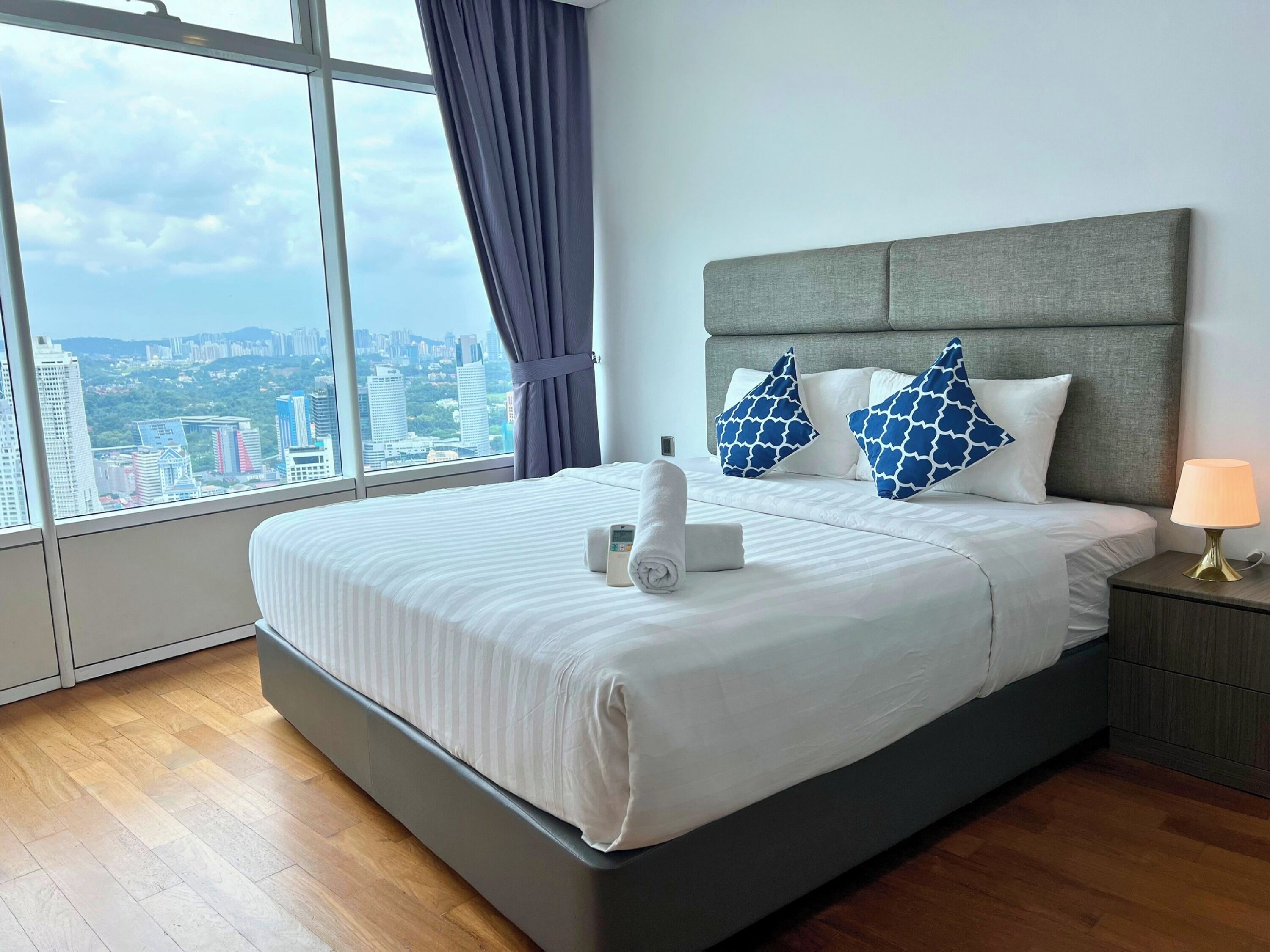 A modern hotel room with a large bed, grey headboard, and a view of the cityscape through large windows.