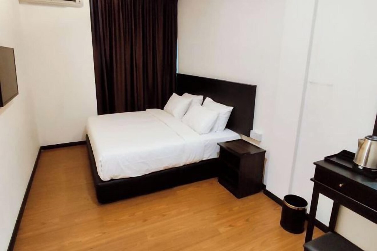 A hotel room with a bed, a nightstand, a small table with a kettle, and a trash can.