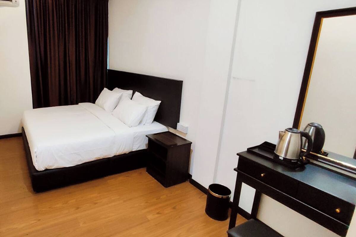 A hotel room with a bed, a nightstand, a small table with a kettle, and a trash can.