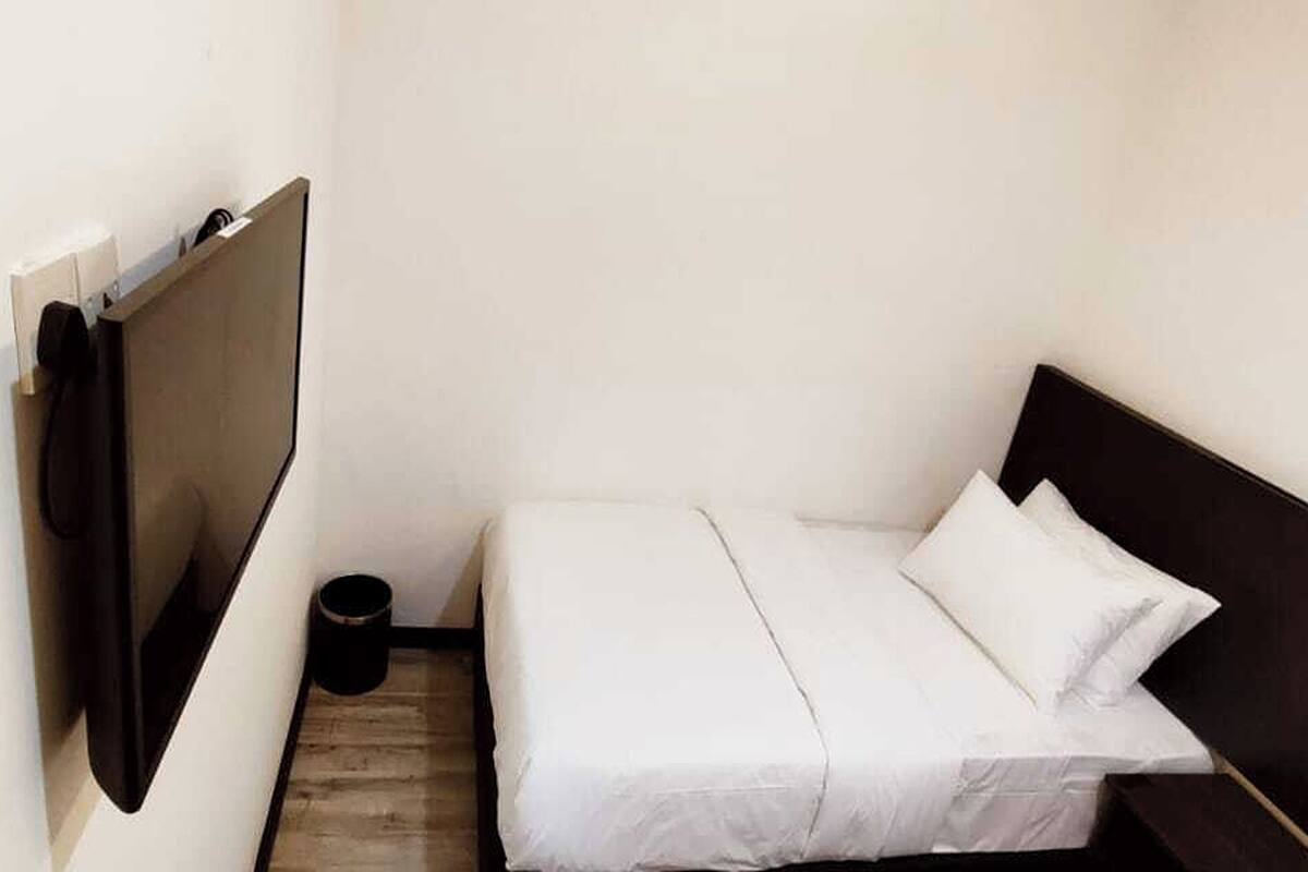 A small, narrow hotel room with a single bed, a wooden floor, and a black trash bin.