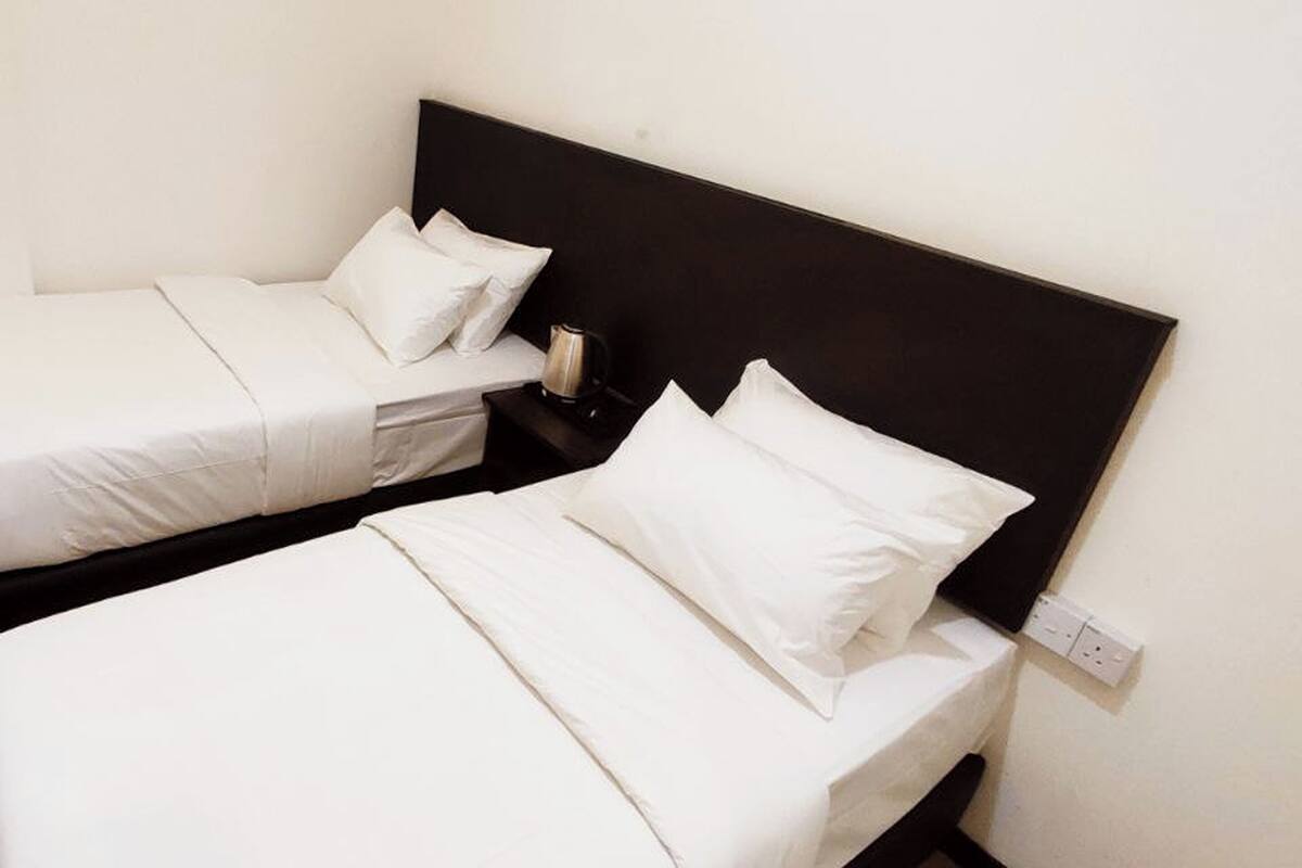 Two single beds with white bedding and a black headboard, a bedside table with a lamp, and a power outlet on the wall.