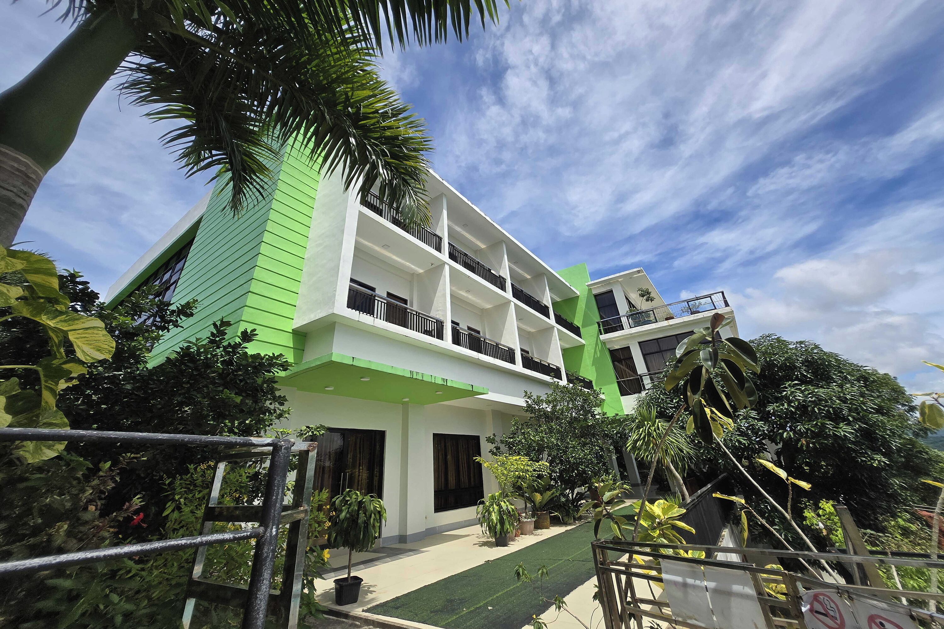 Un edificio moderno de dos pisos con una combinación de colores verde y blanco, rodeado de abundante vegetación y un cielo despejado.