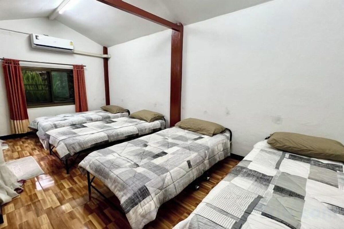A room with two beds, a window with curtains, an air conditioning unit, and a wooden floor.