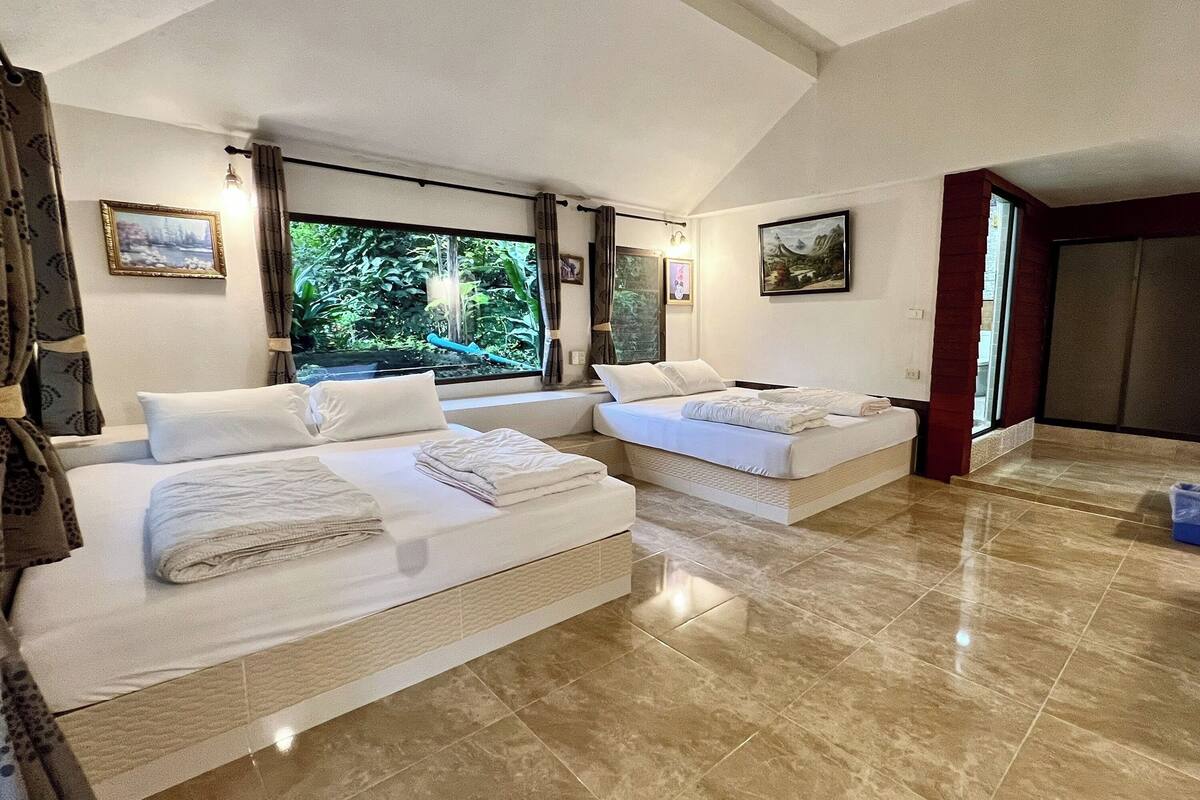 A hotel room with two beds, a large window with a view of greenery, and a tiled floor.