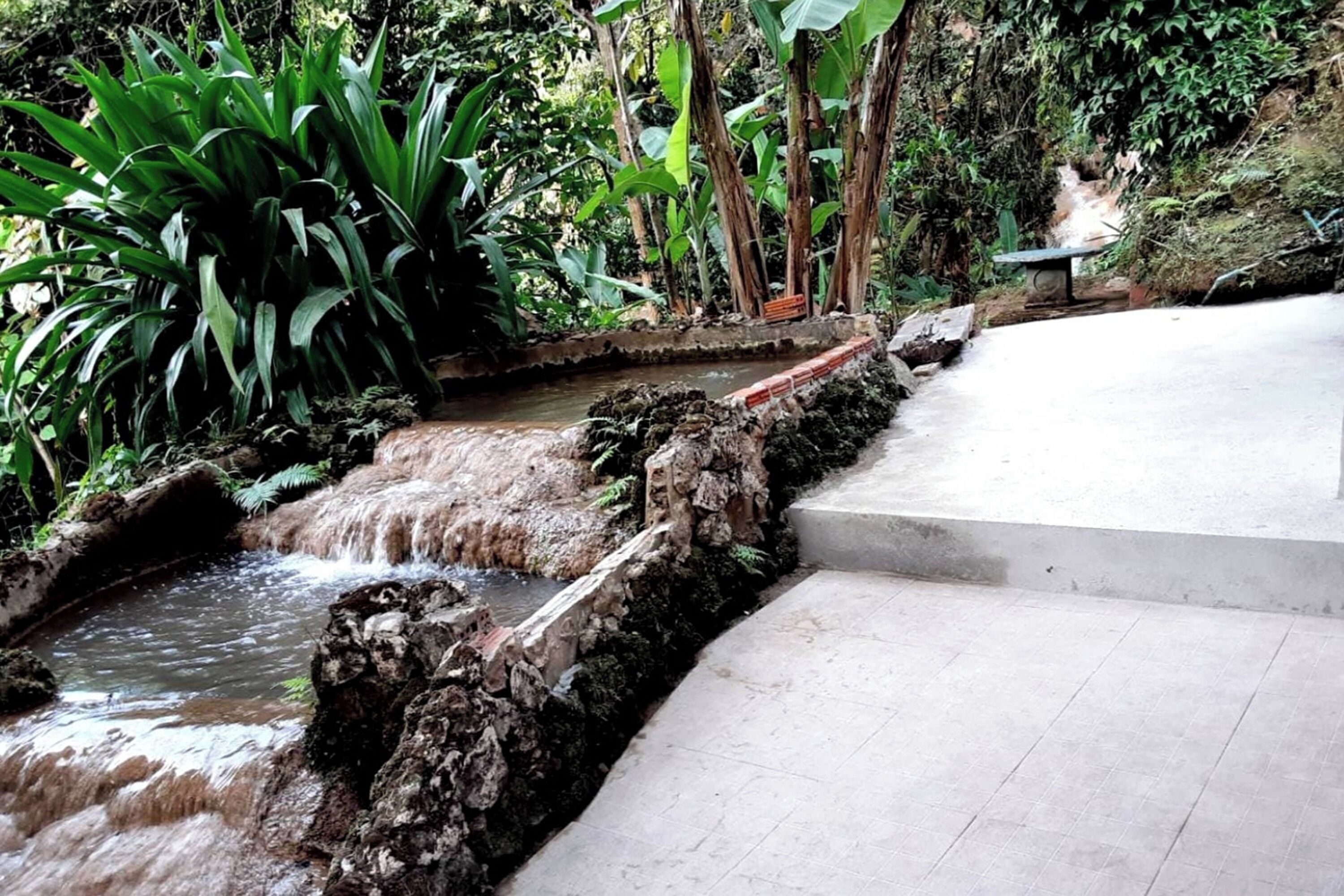 Ein Steinelement mit Wasserspiel und kleinem Wasserfall, umgeben von üppigem Grün und einer Bank.