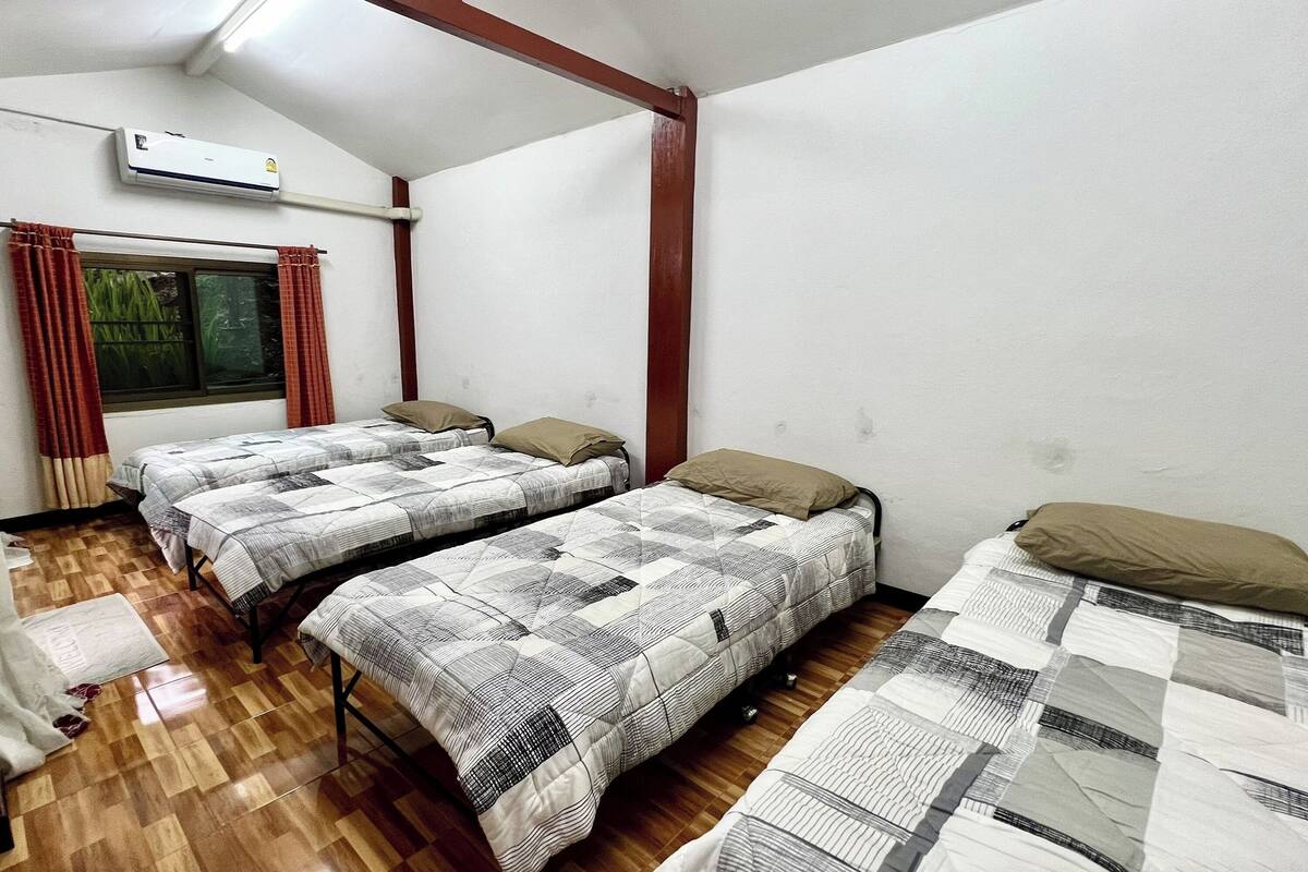 A dormitory room with three beds, wooden flooring, an air conditioning unit, and a window with curtains.