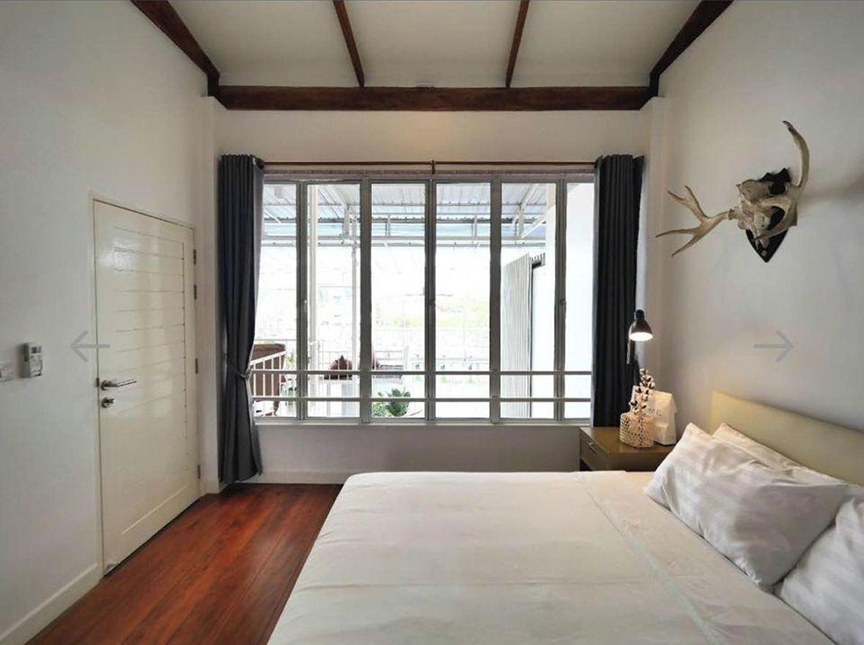 A bedroom with a large bed, a window with curtains, a wooden floor, and a wall-mounted deer skull decoration.