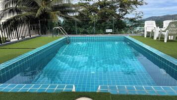 A rectangular swimming pool with blue tiles, surrounded by artificial grass and white plastic chairs.