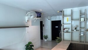 Zona de recepción de hotel con un mostrador de madera, un ventilador blanco, un televisor montado en la pared y una estantería con objetos decorativos.