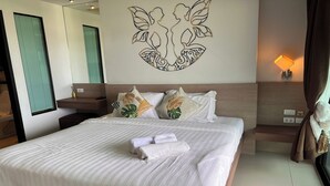 Una cama bien hecha con ropa blanca y toallas enrolladas, una obra de arte en la pared que muestra dos figuras, una mesita de noche con una lámpara y una ventana con persianas.