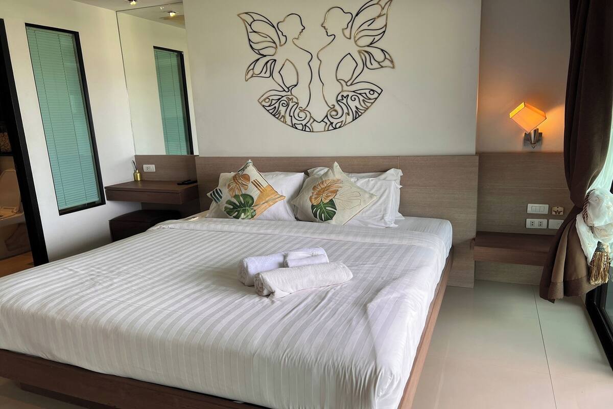 Una cama bien hecha con ropa blanca y toallas enrolladas, una obra de arte en la pared que muestra dos figuras, una mesita de noche con una lámpara y una ventana con persianas.
