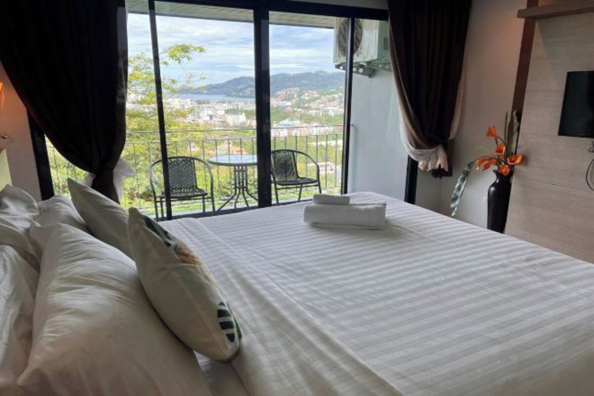 A hotel room with a bed, a small table, and a chair. There is a balcony with a view of a city and mountains.