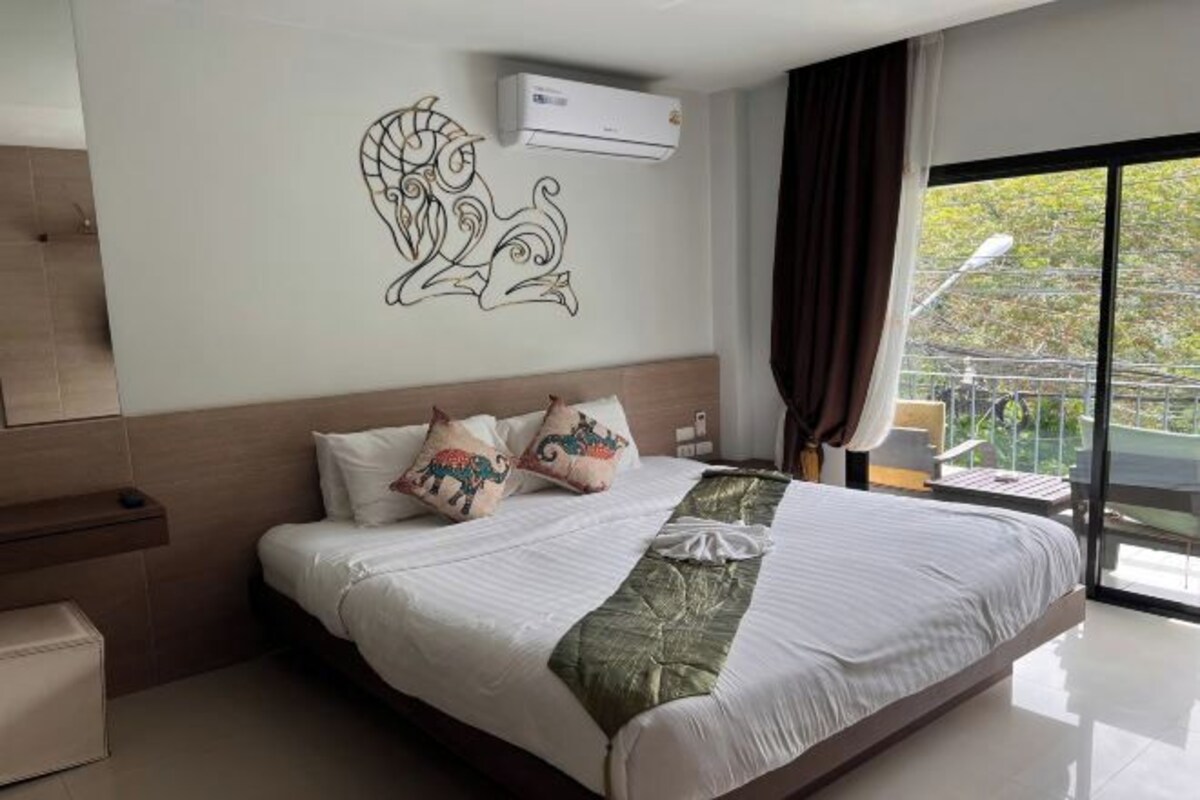 A hotel room with a large bed, a wall-mounted air conditioner, and a view of the outdoors through a large window.