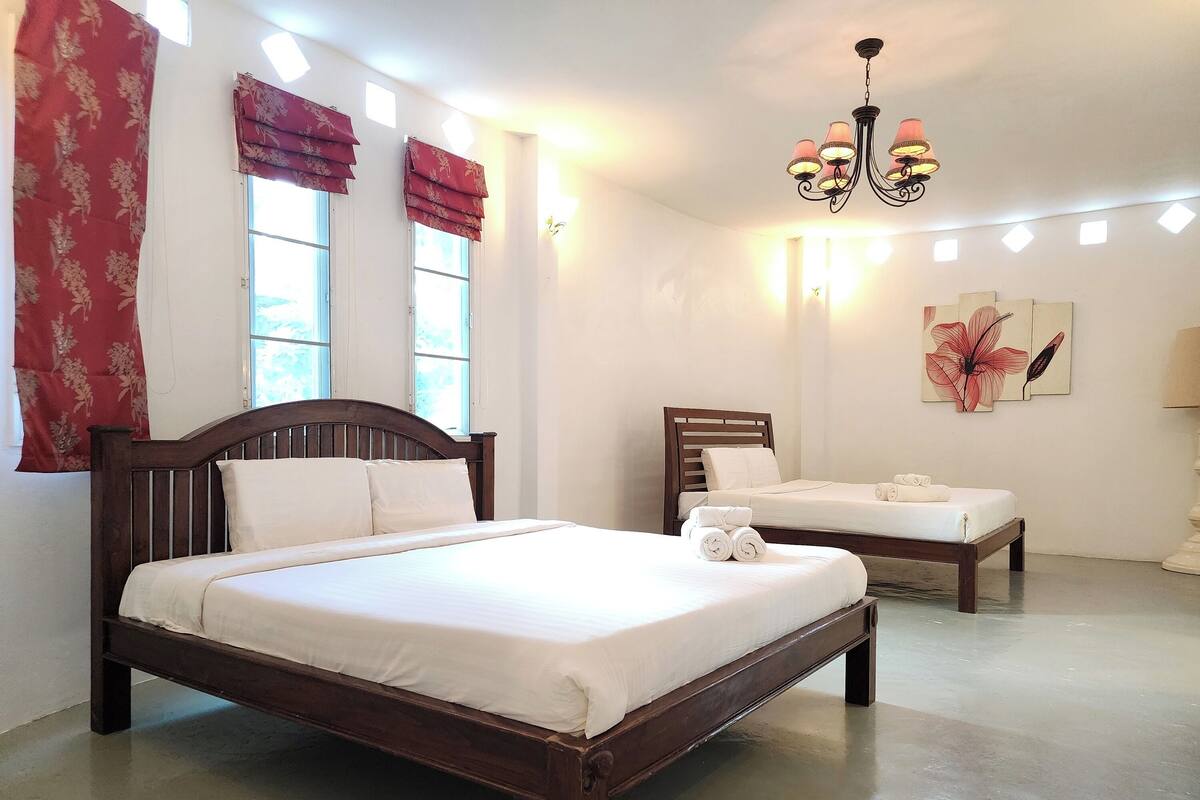 Une chambre d’hôtel avec un grand lit, un lit plus petit et un banc. Il y a des fenêtres avec des rideaux, un lustre et des œuvres d’art accrochées au mur.