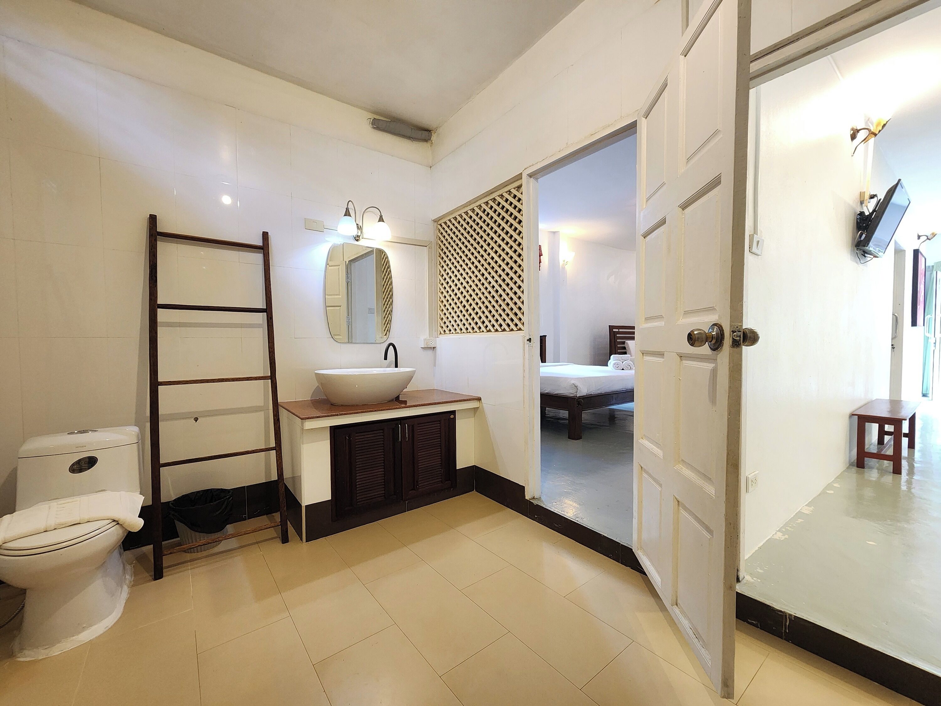 A bathroom with a toilet, sink, mirror, and a bed visible through an open door.
