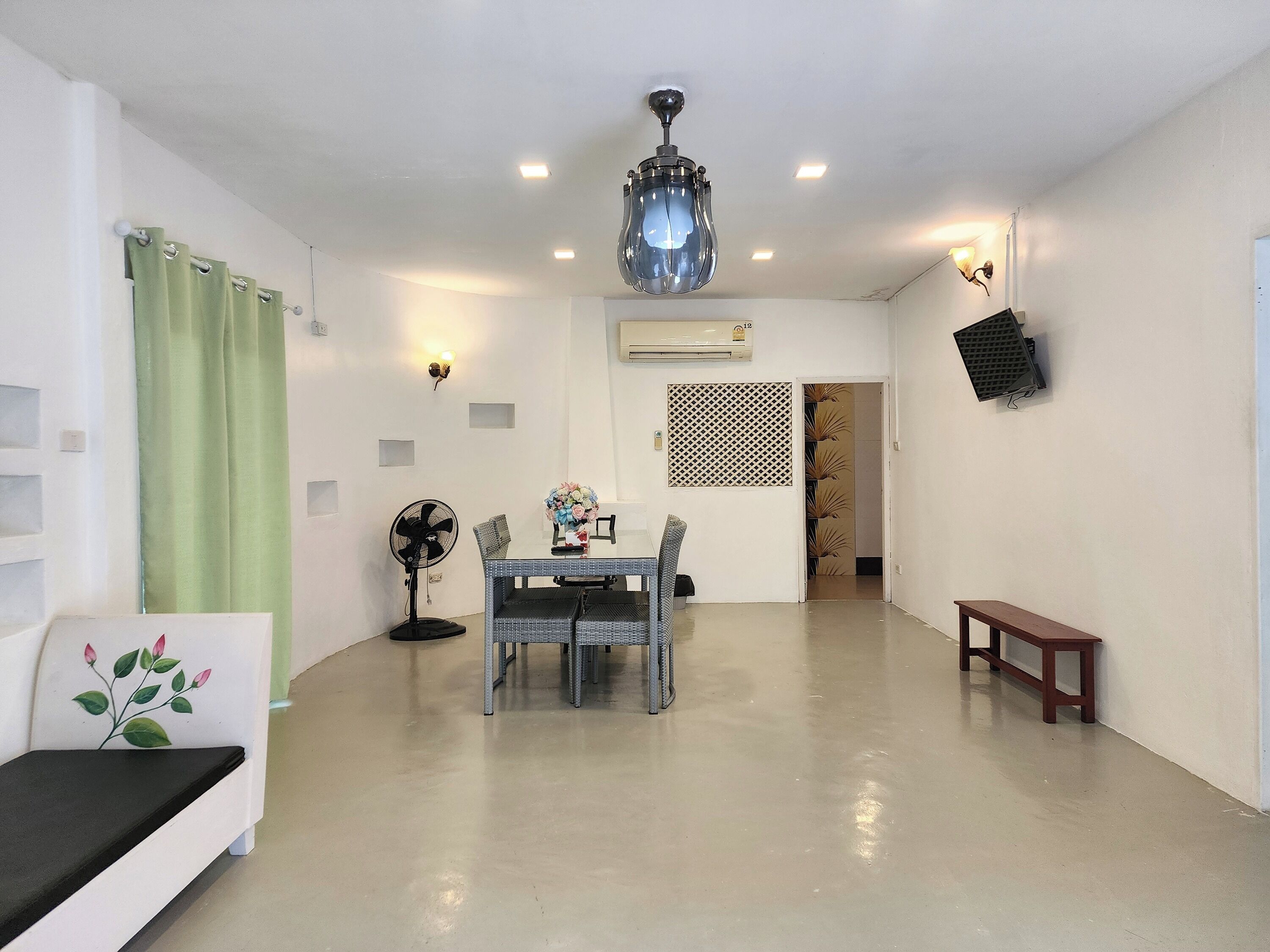 A spacious living area with a dining table, chairs, a sofa, a standing fan, a ceiling fan, and a TV mounted on the wall.