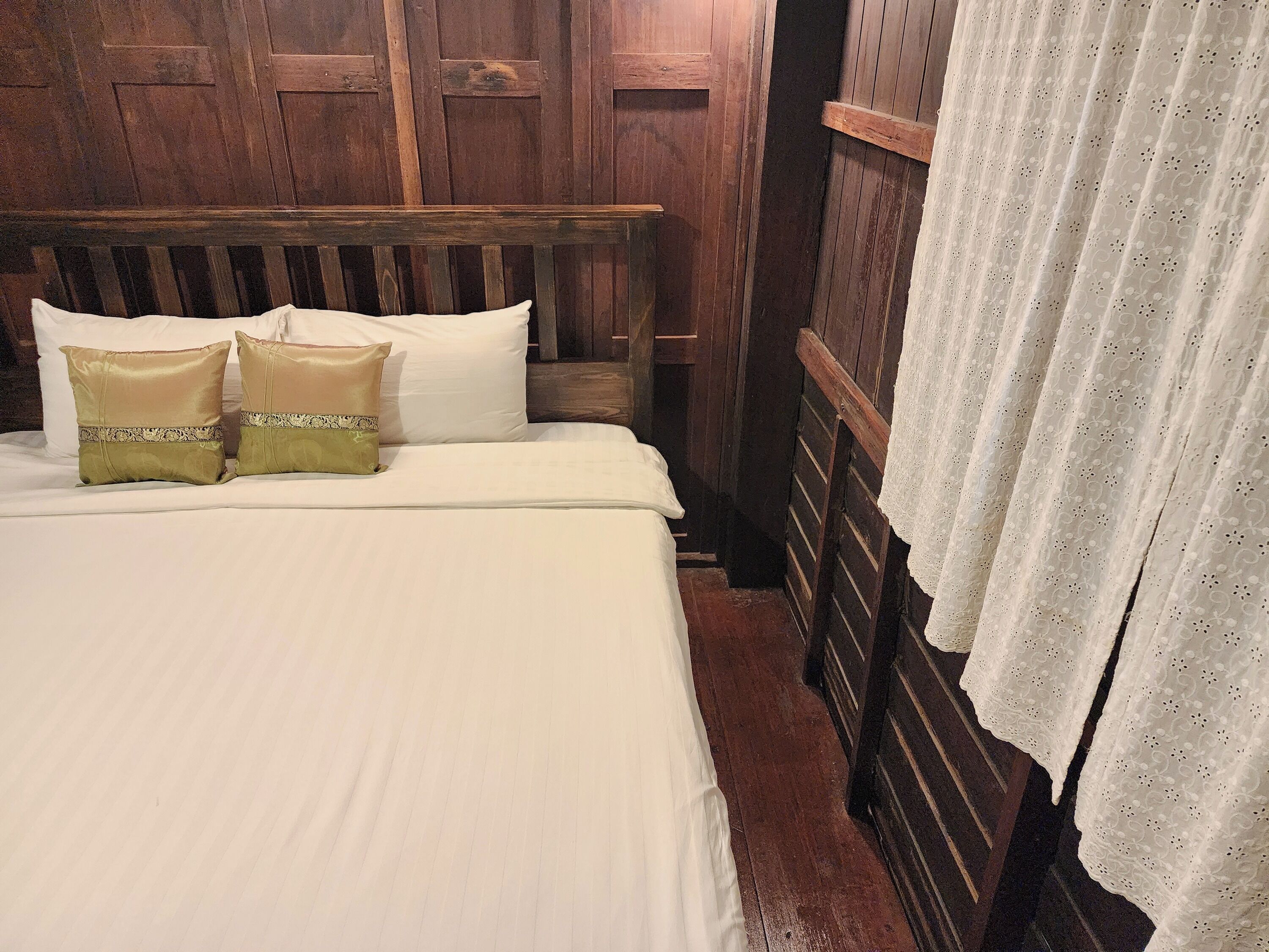Uma cama com roupa de cama branca, duas almofadas decorativas, cabeceira de madeira e cortina de renda.
