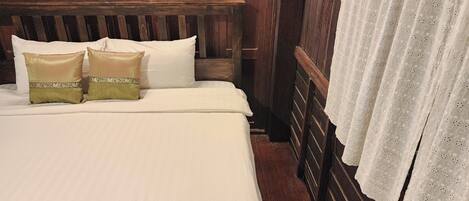 Uma cama com roupa de cama branca, duas almofadas decorativas, cabeceira de madeira e cortina de renda.