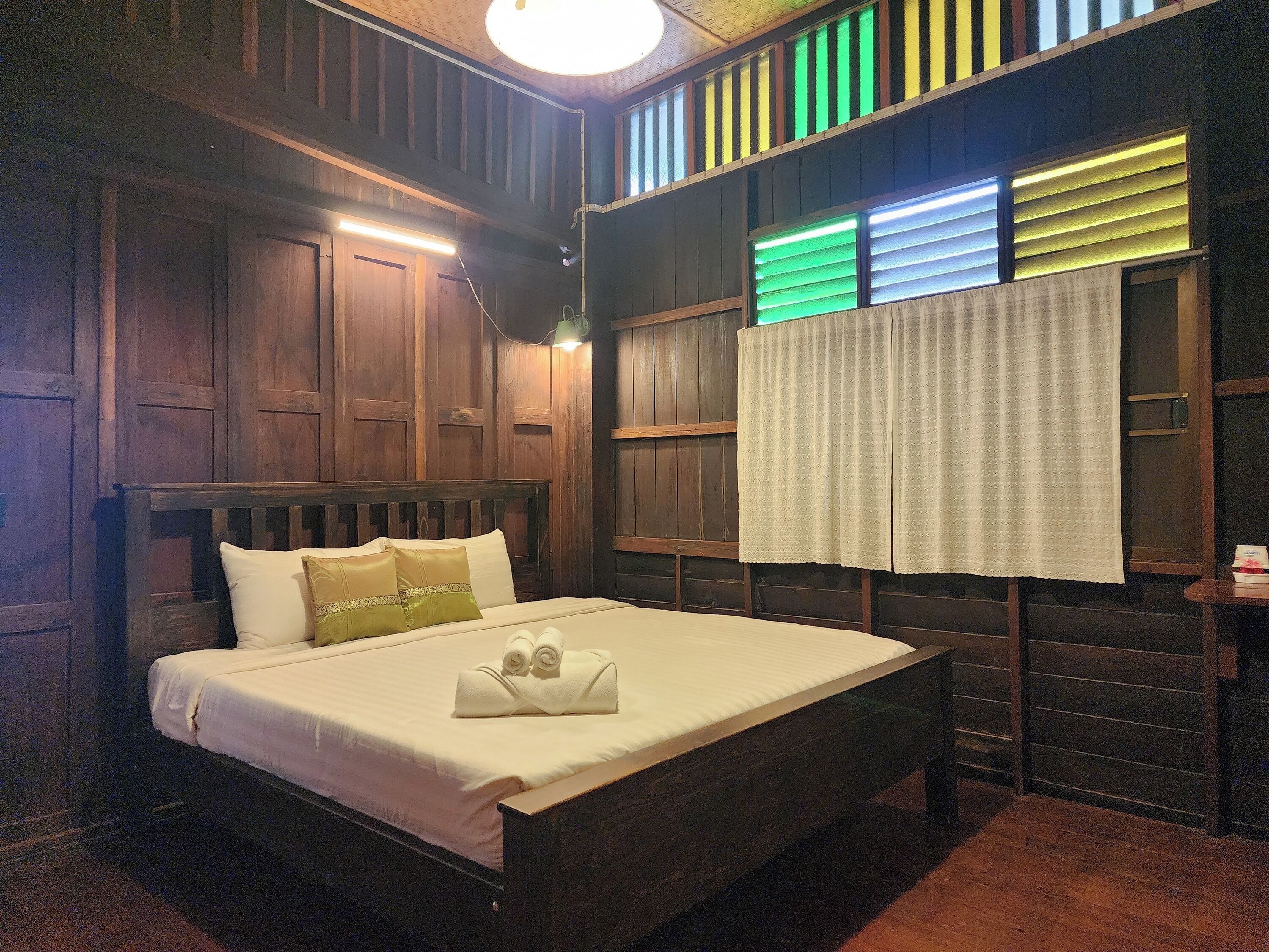 A wooden bed with a white mattress and pillows, a bedside table with a lamp, and a window with green blinds.